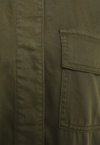 Tissu de veste vert olive avec une surface texturée. Présente une poche à rabat et des détails de coutures visibles. Le matériau semble robuste et durable.