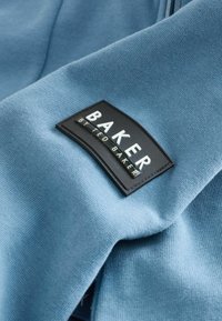 Tessuto azzurro chiaro con una trama liscia, caratterizzato da un'etichetta patch nera con la scritta "BAKER" e "by Ted Baker." Il design è semplice e moderno.