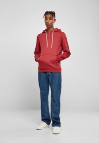 Felpa rossa con cappuccio e lacci bianchi, tasca a marsupio e polsini a costine, abbinata a jeans blu a gamba dritta e sneakers bianche.