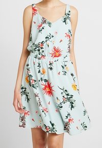 Blommig klänning i ljus mintgrön tyg med V-ringning, justerbara axelband och elastisk midja. Den har livfulla röda och gula blommor.