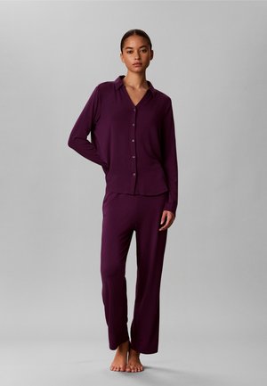 Femme debout pieds nus portant un haut boutonné à manches longues violet foncé assorti et un pantalon ample sur un fond uni gris clair.