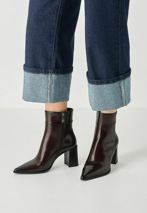 Stivaletti in pelle marrone scuro con punte appuntite e tacchi a blocco, indossati con jeans in denim blu scuro risvoltati.