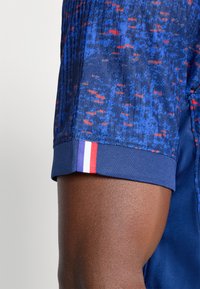 Cămașă cu mânecă scurtă, având un model stropit în nuanțe de albastru și roșu, cu un guler albastru solid și un accent tricolor (albastru, alb, roșu) pe manșetă.