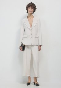 Hellbeige Anzug mit tailliertem Schnitt, einreihiger Jacke, Reverskragen und zwei Vordertaschen; weit geschnittene Hose und schwarze Handtasche.