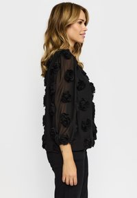 Sort bluse med tredimensionelle stofroser, halvtransparent materiale, lange ærmer og løs pasform. Kombineret med mørke bukser.