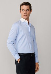 Mann trägt hellblaues Hemd mit weißem Kragen, schwarzen Gürtel und schwarze Hose, steht mit einer Hand in der Tasche und schaut zur Seite.