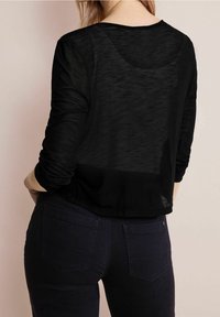 Top negro de manga larga con una tela ligera y transparente. Presenta un escote redondeado y un dobladillo que se sitúa por encima de la cintura, combinado con jeans oscuros.