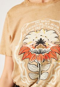 Camiseta de algodón beige con un gran gráfico floral en rojo, blanco y negro, con detalles dorados y texto debajo. Mangas cortas y cuello redondo.