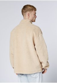 Beige Fleecejacke mit hohem Kragen, sichtbarer weicher Textur und lässiger Passform. Verfügt über angesetzte Schultern und eine entspannte Silhouette.