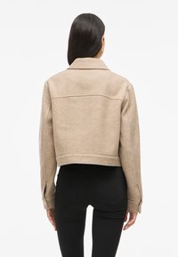 Giacca cropped in lana beige con colletto, caratterizzata da una superficie liscia e cuciture sulle spalle. Indossata sopra pantaloni neri, vista da dietro.