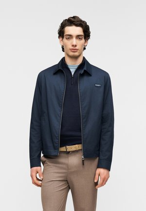 Veste zippée bleu marine avec col et écusson logo, portée sur un pull sombre, associée à un pantalon beige et une ceinture. Texture de tissu lisse.