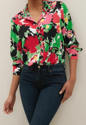 Femme portant une chemise fleurie colorée à boutons avec des motifs rouges, verts, noirs et roses, associée à un jean bleu foncé.