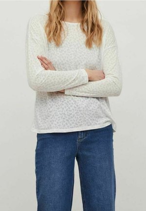 Personne aux longs cheveux blonds portant une chemise légère à manches longues imprimée léopard et un jean bleu, bras croisés devant un fond uni.