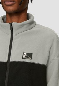 Fleecejas in lichtgrijs en zwart, hoge kraag, ritssluiting, met een vierkant logopatch op de borst en zichtbare stikdetails.