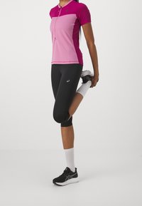 Camisa deportiva ajustada de manga corta en rosa y púrpura con cremallera, combinada con leggings cortos negros. Zapatillas de correr negras y calcetines blancos.