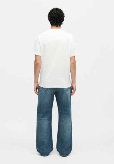 Άνδρας που στέκεται με την πλάτη, φοράει ένα απλό λευκό μπλουζάκι και φαρδιά μπλε τζιν, σε λευκό φόντο.
