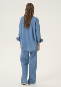 Světle modrá denimová košile s oversized střihem, spadlými rameny a knoflíkovými manžetami. Spárována s volnými denimovými kalhotami ve stejném stylu.