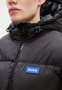 Μαύρο γεμιστό μπουφάν με υψηλό γιακά, κλείσιμο φερμουάρ, κορδόνια και μπλε λάβαρο "HUGO". Απαλή υφή υφάσματος και ραμμένες λεπτομέρειες.