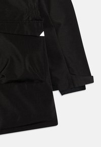 Blízký záběr na rukáv a kapsu černé bundy, který ukazuje elastické nařasení a detaily manžety na jednobarevném světle šedém pozadí.