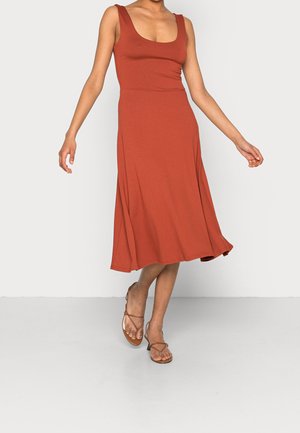 Femme portant une robe midi sans manches rouge rouille et des sandales marron à talons avec des brides, debout les bras doucement étendus.