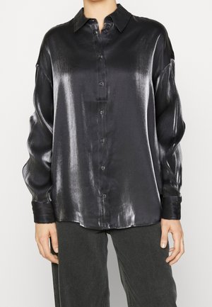 Kobieta ubrana w błyszczącą czarną satynową koszulę z długim rękawem i zapięciem na guziki, z kołnierzykiem, oraz ciemne spodnie, stojąca na jasnym tle.