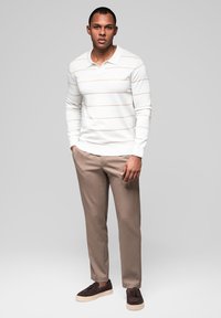 Muž stojící, oblečený v bílé košili s dlouhým rukávem a světlými horizontálními pruhy, béžových kalhotách a tmavě hnědých slip-on botách.