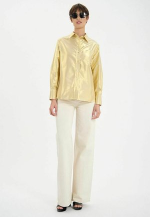 Chemise en satin doré à manches longues avec un devant à bouton et une poche poitrine, associée à un pantalon blanc évasé et des sandales à talons noires.