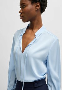 Blusa a maniche lunghe azzurro chiaro con scollatura a V e lacci al collo, caratterizzata da un tessuto morbido e una vestibilità rilassata.