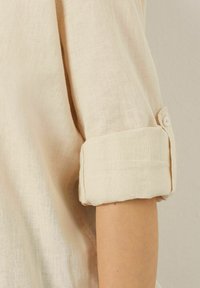 Camisa de lino beige con mangas remangadas. La textura parece ligera y ligeramente arrugada, con un detalle de botón en el puño.