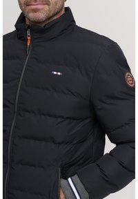 Svart dunjakke med glidelås foran, med quiltet tekstur, horisontale sømmer og oransje detaljer. Logomerke på venstre erme.