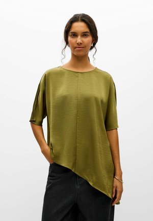 Une femme pose sur fond blanc, vêtue d’un chemisier asymétrique vert olive à manches courtes et d’un jean foncé.
