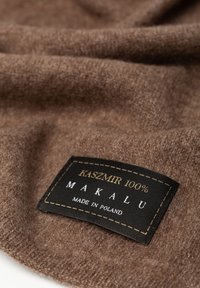 Brązowy kaszmirowy materiał o miękkiej fakturze, z czarną metką, na której widnieje napis "KASZMIR 100% MAKALU MADE IN POLAND" w białym i złotym stylu.