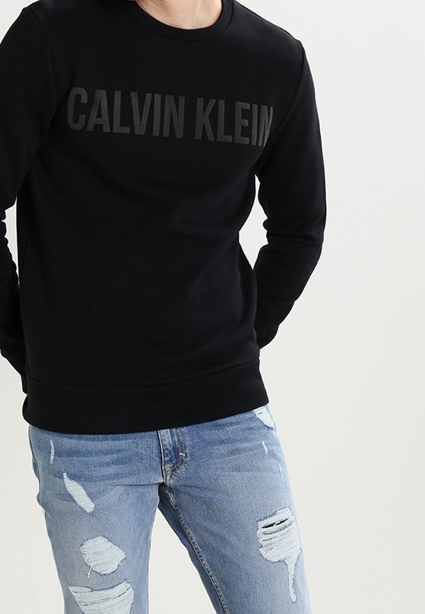 Personne portant un sweat-shirt noir Calvin Klein et un jean bleu clair déchiré, debout devant un fond clair uni.