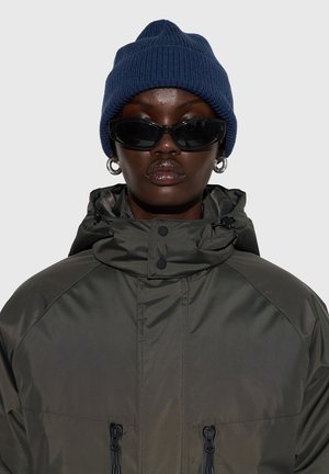 Dark green puffer jacket with a high collar, featuring drawstrings and zipper details. Accompanied by a blue knit beanie and black sunglasses.