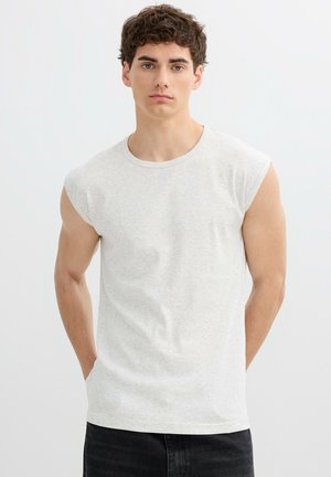 Joven con pelo rizado que lleva una camiseta sin mangas gris claro y pantalones oscuros, de pie con las manos detrás de la espalda sobre un fondo blanco.