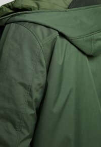 Chaqueta verde con capucha vista desde atrás. Fabricada en tela mate con detalles de costura. Presenta una capucha redondeada y una textura lisa.