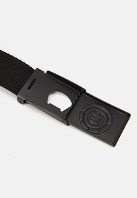 Cinturón de nylon negro con una hebilla de metal texturizada que presenta un logo circular y un corte. El cinturón está tejido para mayor durabilidad y flexibilidad.