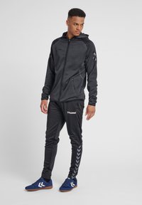 Zwarte rits-hoodie van textuurstof, met een aansluitend ontwerp en capuchon; gecombineerd met zwarte joggerbroeken met zijstrepen en logo-accenten. Blauwe sportschoenen.