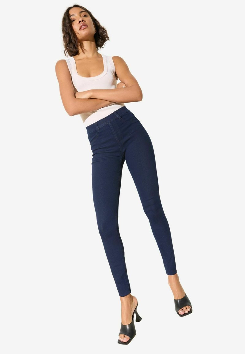 Tumesinised kõrge vöökohaga kitsad teksapüksid ühtlase tekstuuriga, mida kannab kreemikas valge ümarakaelusega topp ja mustad avatud varbaosaga kontsakingad.