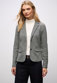 Blonde vrouw die een grijze blazer draagt over een witte coltrui en donkere jeans, staand tegen een effen lichte achtergrond.