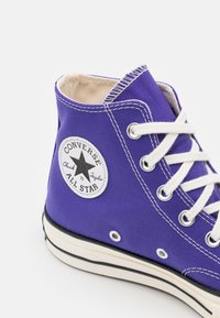 Fioletowy płócienny sneakers z wysoką cholewką, z białymi oczkami i sznurówkami, gumową podeszwą i czarnym paskiem, z okrągłą naszywką z logo po boku.