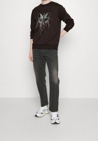 Sudadera marrón con gráfico de mariposa y rayo, combinada con vaqueros gris oscuro y zapatillas blancas gruesas. Ajuste casual y relajado.