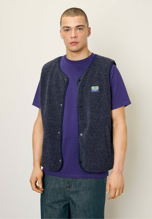 Gilet en polaire marine avec boutons-pression, doté de poches latérales et d'une patch logo. Porté sur une chemise violette, associé à un jean.