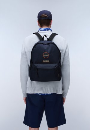 Mand iført navyblå rygsæk, lysegrå sweater, navy shorts og navy kasket, set bagfra mod en ensartet lys baggrund.