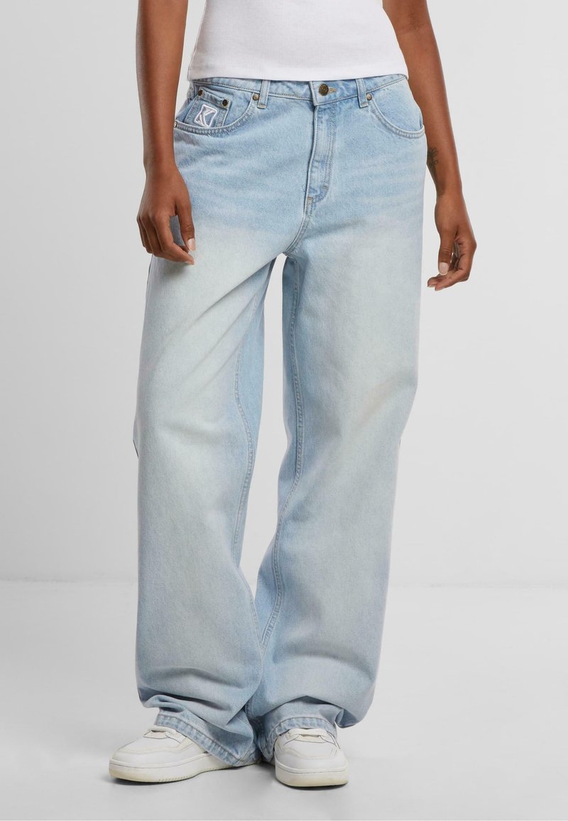 Personne portant un jean denim bleu clair ample, des baskets blanches et un haut blanc sans manches, debout devant un fond uni.