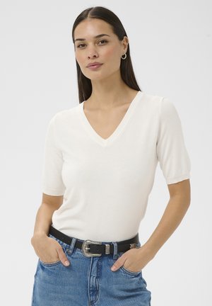 Frau mit langen dunklen Haaren, die ein weißes V-Ausschnitt-Kurzarmshirt und blaue Jeans mit schwarzem Gürtel trägt, Hände in den Taschen, neutraler Hintergrund.