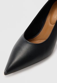 Sort læder højhælet sko med spidse tæer, glat tekstur og en udsparing på siden, der afslører en tan indersål.
