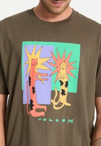 Oliven grøn bomulds T-shirt med et farverigt grafik af to stiliserede katte med solstråle accenter, omridset i sort. Logo tekst nedenfor.