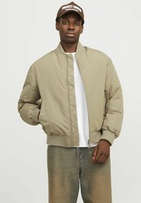 Veste bomber beige avec un motif matelassé, des poignets côtelés, une fermeture éclair sur le devant et des poches latérales. Associée à une chemise blanche et un jean en denim.