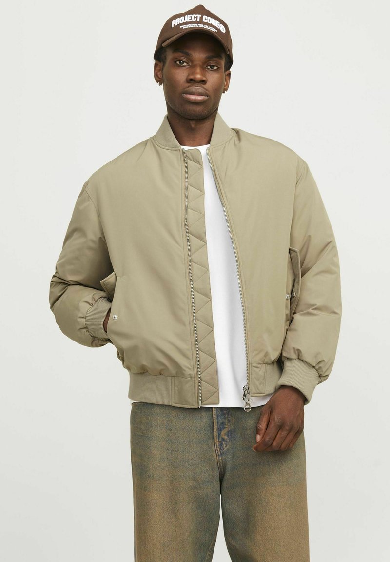 Veste bomber beige avec un motif matelassé, des poignets côtelés, une fermeture éclair sur le devant et des poches latérales. Associée à une chemise blanche et un jean en denim.